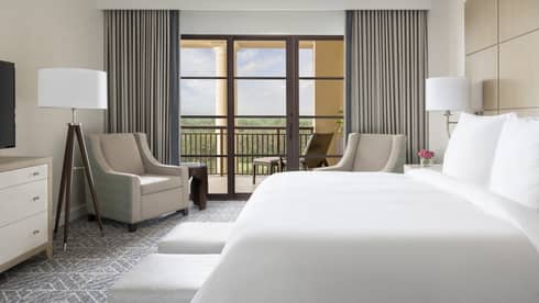 Hotel room with white king bed, two beige arm chairs, grey curtains, walk-out balcony