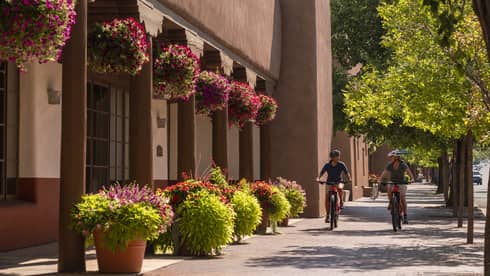 Two people on bikes riding down a small street with a building on one side and small trees on the other.