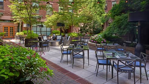 An outdoor eating area with metal chairs and glass tables surrounded by plants.