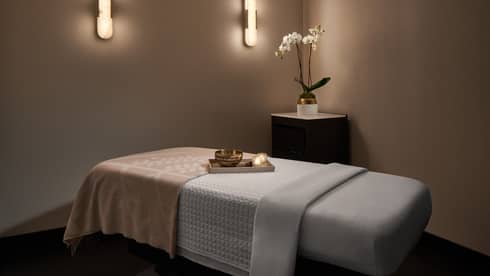 A softly lit spa treatment room in warm neutral colours features a treatment table with sheets and a blanket, mellow wall sconce lighting, and a corner table with a floral arrangement.