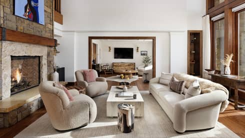 Mountain residence living room with a stone fireplace, sofa and armchairs around a coffee table, leading into a connecting seating area with a TV on the wall