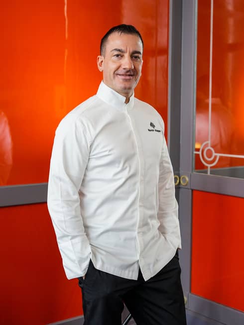 A smiling Chef Marco Violano in a white chef’s jacket casually posing, with hands in pockets, in front of a red wall