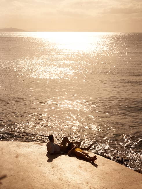 Two people lounge on stone terrace right next to the sea at sunset