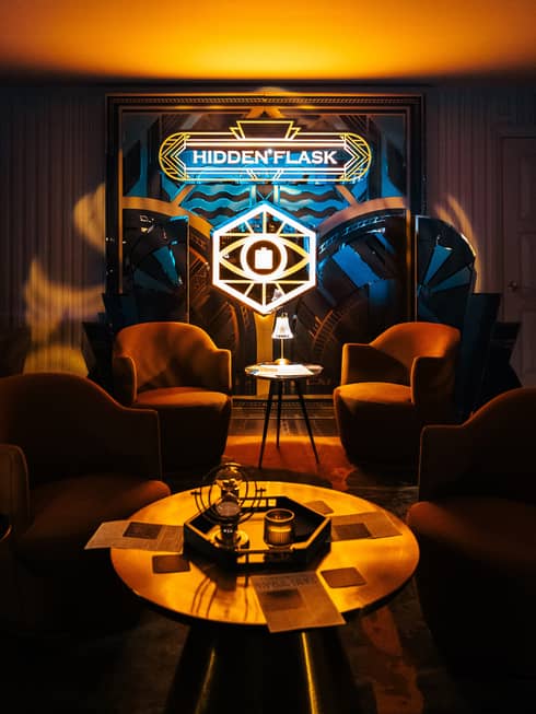 Dimly lit lounge area with four arm chairs, a round coffee table and a sign that says "Hidden Flask"
