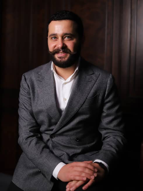 Man with short dark hair and trimmed beard wearing a grey suite with a white collared shirt