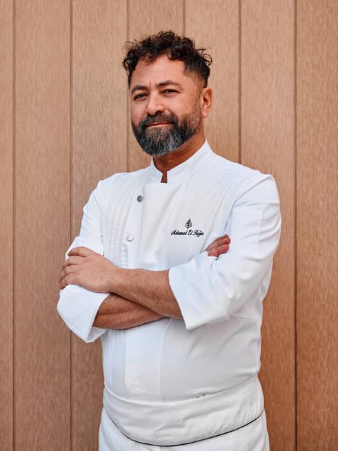 Chef with short dark hair and a trimmed beard stands wearing a white-long-sleeved chef's jacket and apron in front of a light-coloured wall
