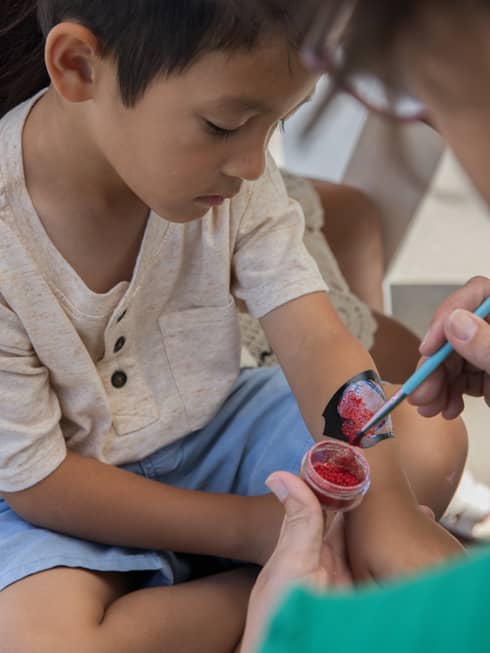 Child wearing beige shirt and light blue shorts sits getting their arm painted