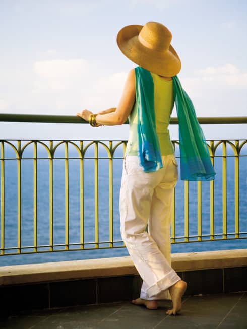 Back view of woman wearing sun hat, flowing blue-and-green scarf, standing at patio railing looking at sea