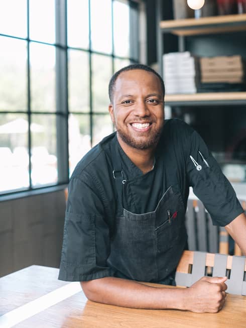 Portrait photo of Chef de Cuisine James Waters wearing black uniform, smiling in front of window