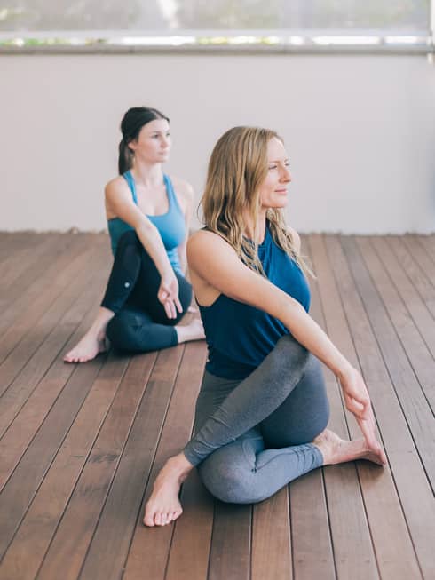 One blonde woman and one brunette woman sit with one leg crossed over the other with their bodies turned to one side and their front hands forming a lam mudra
