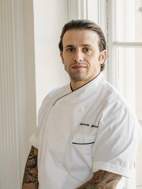 A chef standing near a window wearing a white chef's coat.
