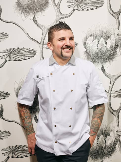 A chef with arm tattoos standing in front of a wall with flower wallpaper.