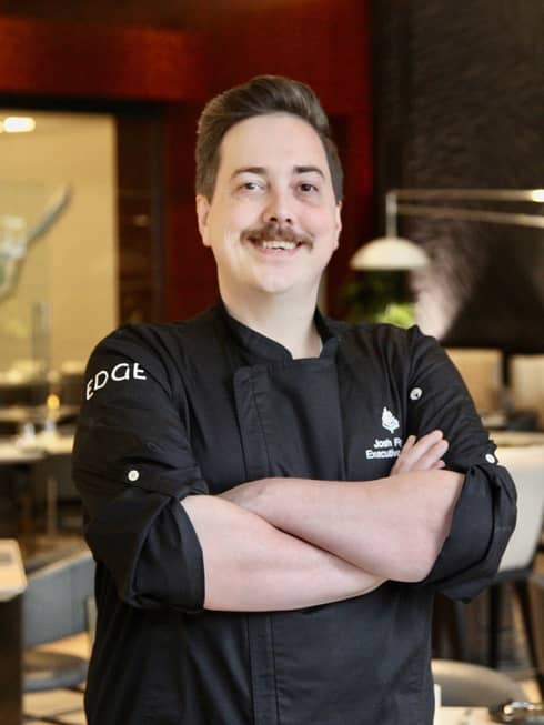 A chef in a black uniform stands with arms crossed in an elegant indoor dining space