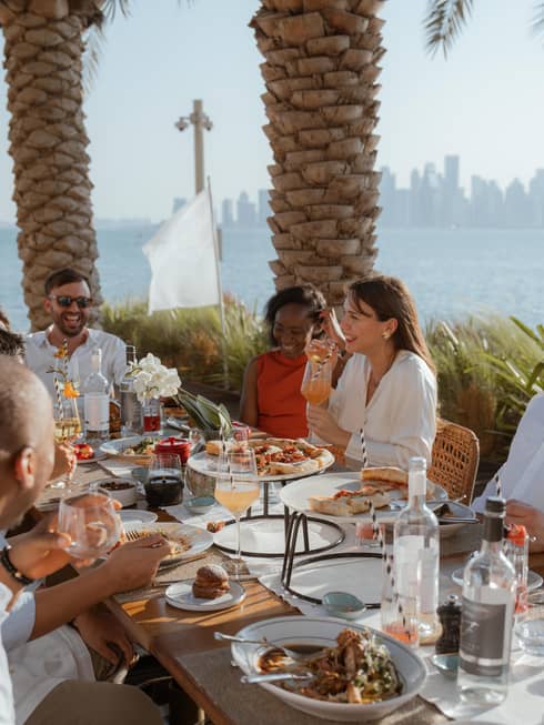 Group sits around long outdoor dining table eating brunch with the ocean and a city skyline in the background