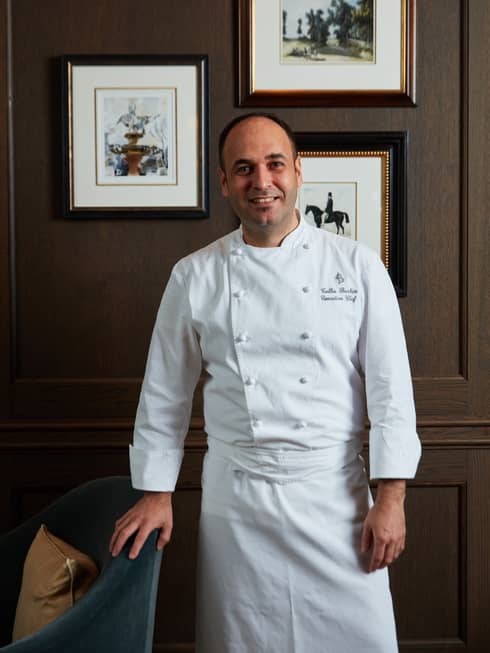 A chef standing in front of framed art and a dark wood wall.