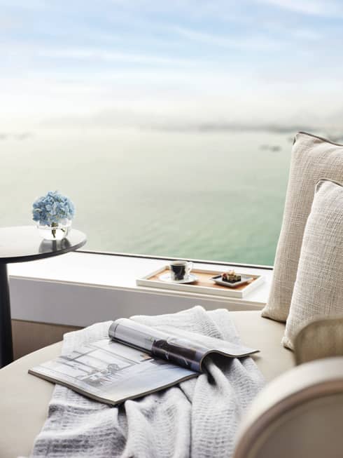 Close-up of cozy seating by window with a cup of coffee, pastries on a tray, a magazine, and blue flowers, overlooking water.