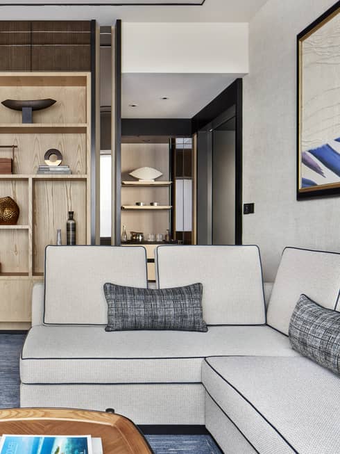 Modern living room with light gray sectional sofa, gray accent pillow, wooden shelves with decor and a view of a small open kitchen area.