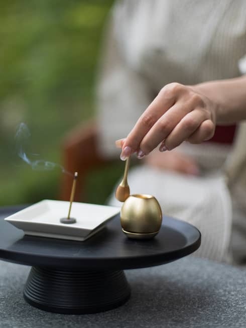 Close-up of a hand holding a small brass spoon over an orin bell and incense burner, with lit incense stick emitting smoke