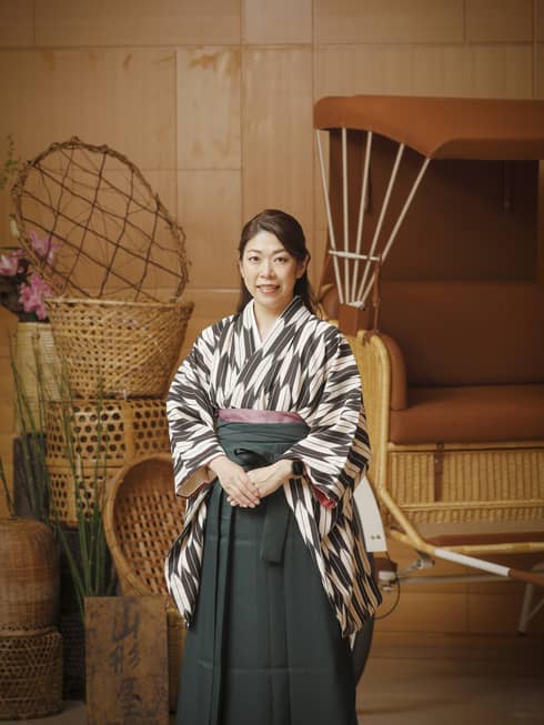 Person in traditional Japanese dress stands in front of pile of woven baskets and rickshaw