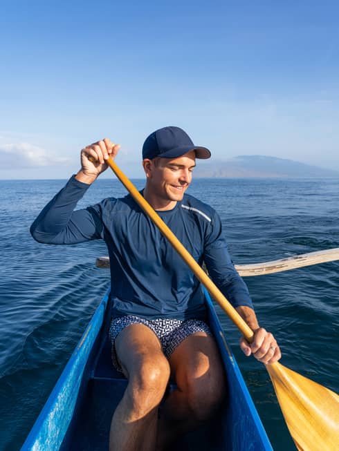 A smiling Gray Malin holds a paddle in a canoe in the open ocean, the East Maui Volcano looming in the background