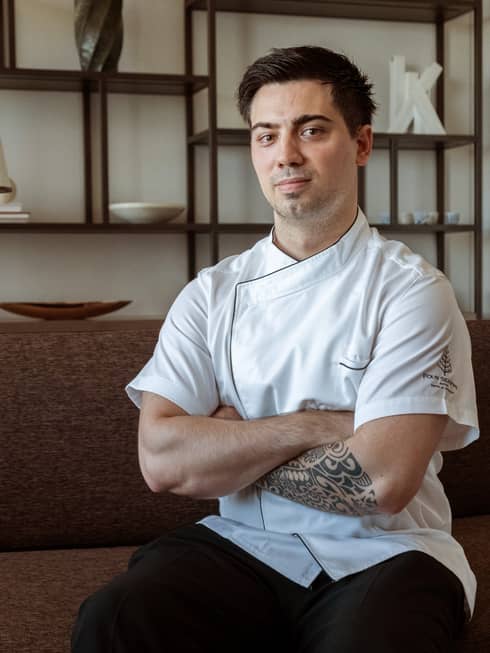 Pizza specialist DeLeo wearing white chef's coat sits on couch with arms crossed