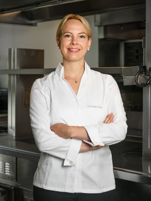 A woman in business attire with blonde hair is smiling and standing in front of a kitchen.