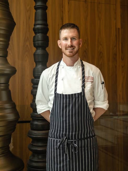 Chef de Cuisine Colin Henderson stands wearing a black-and-white striped apron over a white chef's coat