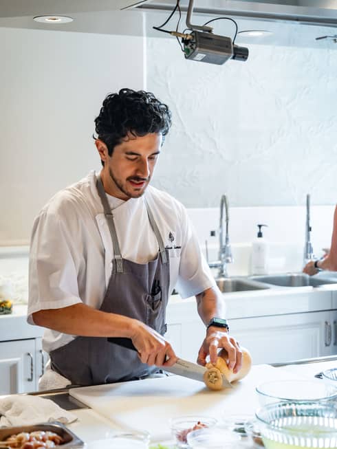 Chef wearing white short-sleeve button-up shirt and a slate grey apron slices squash on a white marble countertop