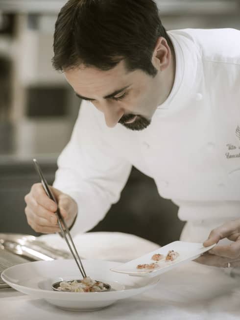 Executive Chef Vito Mollica in white uniform, picks up small shrimp with kitchen tweezers