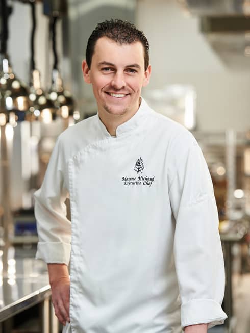 A male chef in chef clothes smiling in a well lit kitchen.