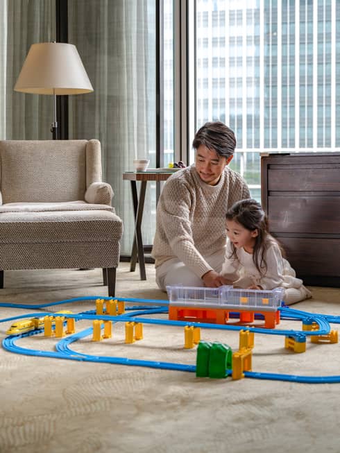 Man and young girl play with toy train set in guest room living area