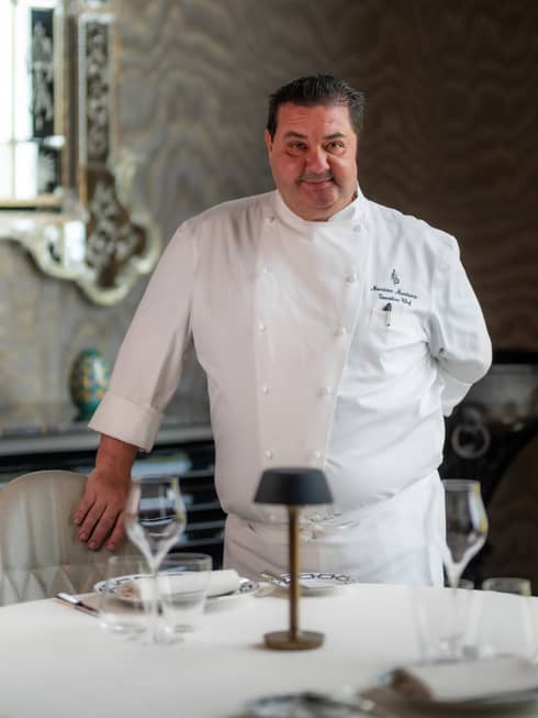 Person in double-breasted white chef?s coat stands behind table with hand on chair