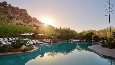 Pool with lounge chairs and umbrellas and the sun setting in the background.