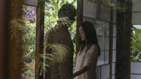 In a glass hut surrounded by abundant greenery, a couple stands at the open entrance. One places a hand on the other’s arm. 
