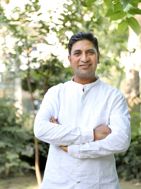 Smiling person in long-sleeved white linen shirt stands outdoors with arms crossed