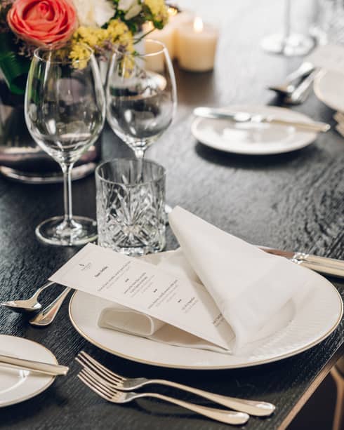 An elegant place-setting on a table for an event, including menu card, napkin, utensils, glassware, dishware, and a floral centrepiece and candles