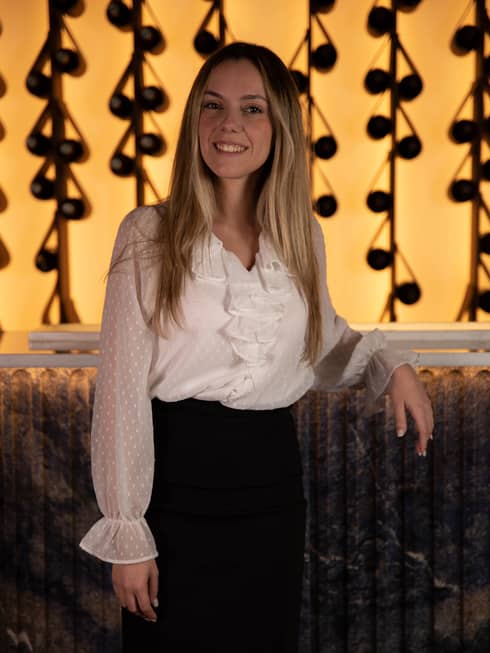Monica Duarte in white ruffled blouse and black pencil skirt in front of gleaming restaurant wall