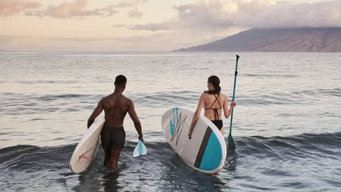 Two people standing in the ocean holding paddleboards with mountains in the background