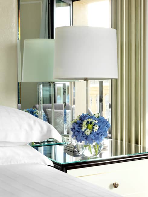 Close-up of corner of bed with white linens, nightstand with white lamp, purple flowers, mirror