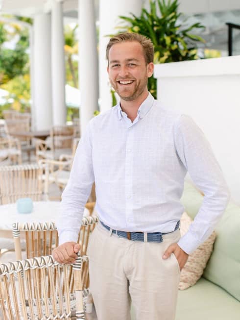 A man in a light blue collared shirt leaning on a chair in a dining area outside.