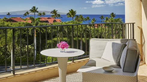 Resort balcony with round table holding pink flowers, cushioned seating and view of tropical landscape and ocean