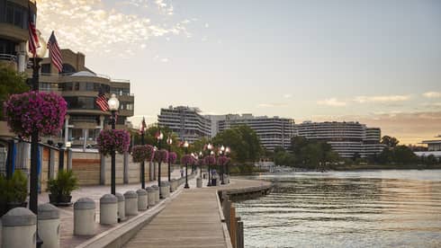 A waterfront with a wooden walkway next to light posts and flowers with building on the other side of a small road.