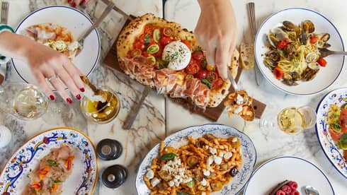 A marble table features white wine and an array of colourful Mediterranean delicacies, hands reaching in to sample the food.