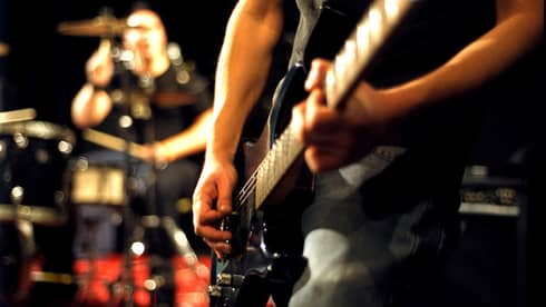 Close-up of musician's arms and hands playing an electric guitar with drumset in the background.