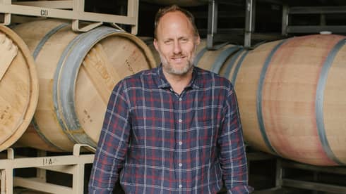 Thomas Brown stands before two racks of wine barrels as he rests his hands on two upright barrels and smiles at the camera.
