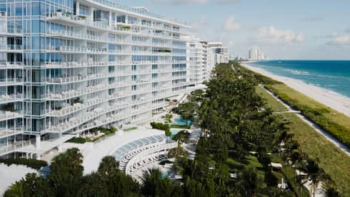 Luxury beachfront hotel with modern glass architecture, lush greenery, a pool area and a view of the ocean and sandy beach under a blue sky