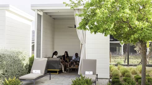Three guests eat snacks on a large couch in a square cabana with a tied-off curtain and two lounge chairs at the entrance.