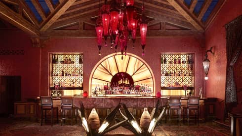 Indoor bar with illuminated shelves holding bottles, red hanging light fixture, geometric ceiling