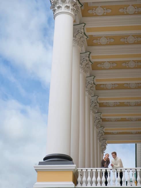 Looking up at man and woman peering over white corner balcony under tall white pillars, gold ornate ceiling