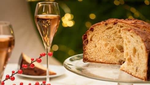 Loaf of panetonne sits on a crystal cake stand with two slices plated on the table next to it, two glasses of pink-hued wine in champagne flutes and a cup of tea
